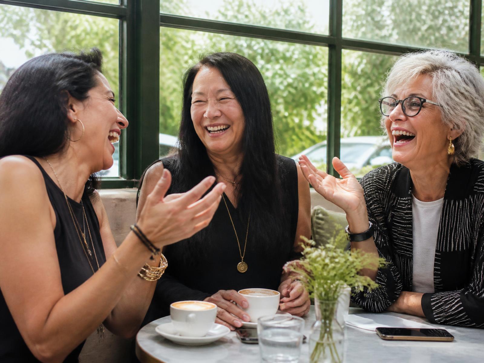 Three women of different ages laughing together over coffee at a sunny café